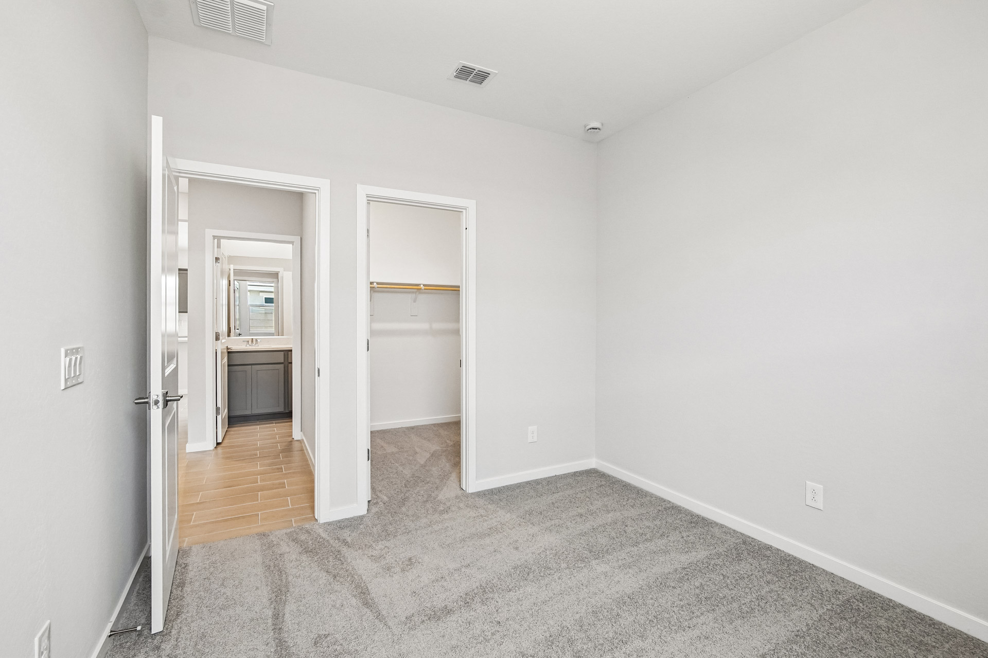 Empty bedroom with light gray carpet, white walls, and two doors; one opens to a bathroom with a vanity, the other to a walk-in closet with shelves.