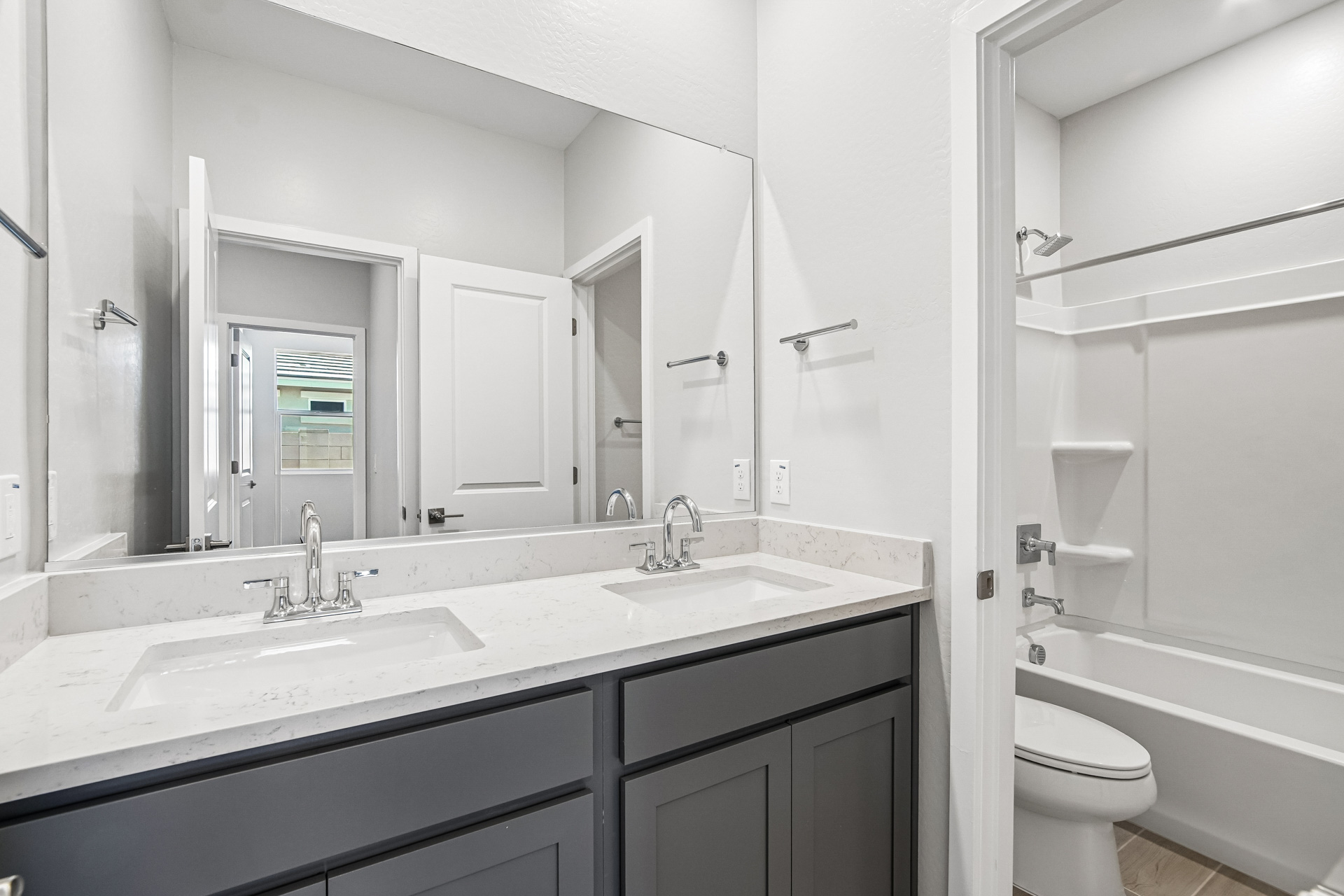 Modern bathroom with double sink vanity, large mirror, gray cabinets, white countertop, and adjacent bathtub with shower and toilet in a small, bright space.