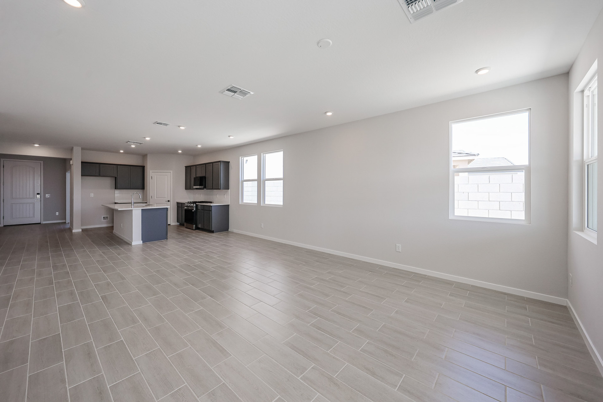 Spacious empty open-plan living area with beige tile flooring, white walls, large windows, and a modern kitchen in the background.