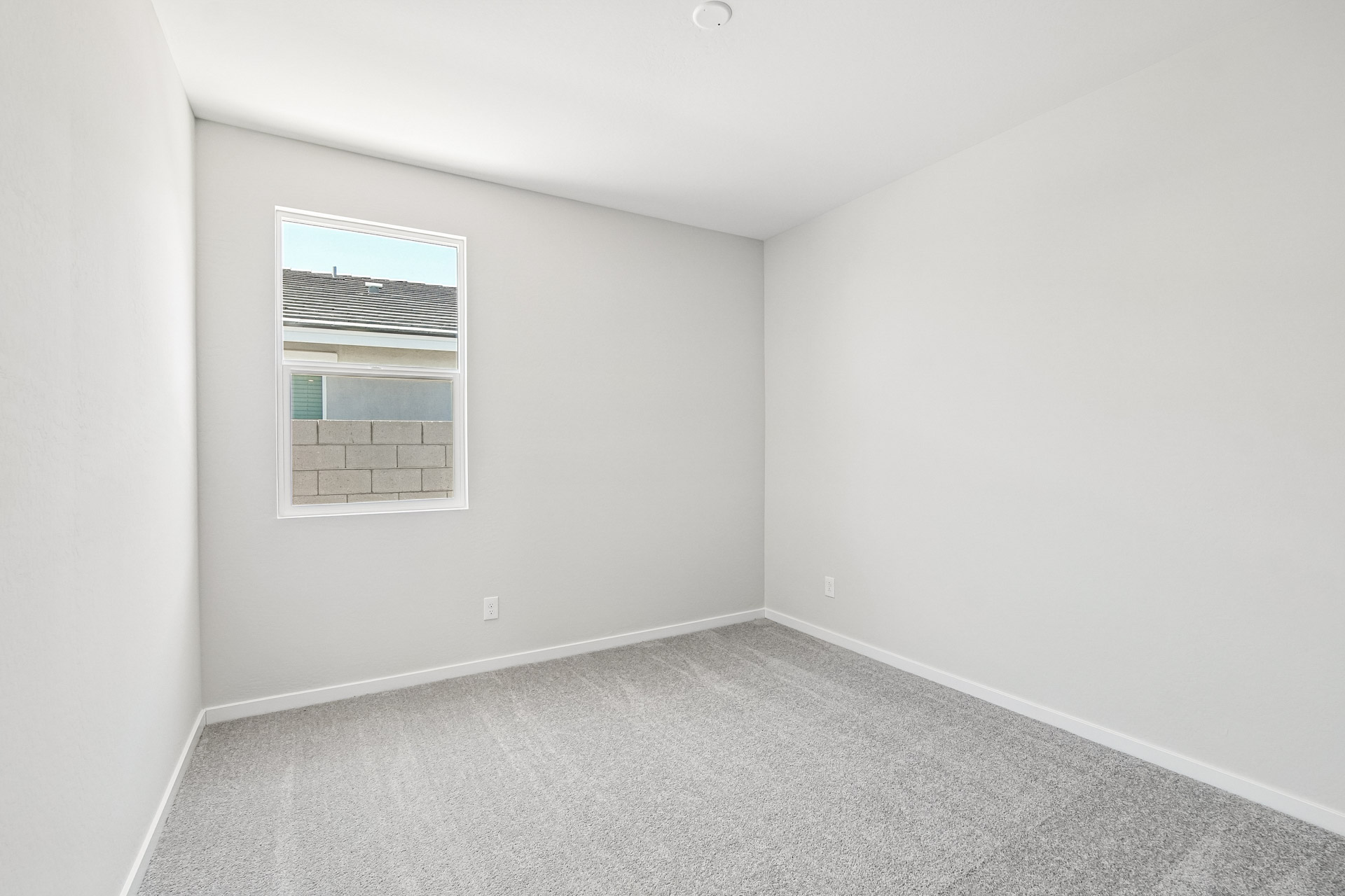 Empty room with light gray carpet, white walls, and a single window showing an outside brick wall and part of a roof.