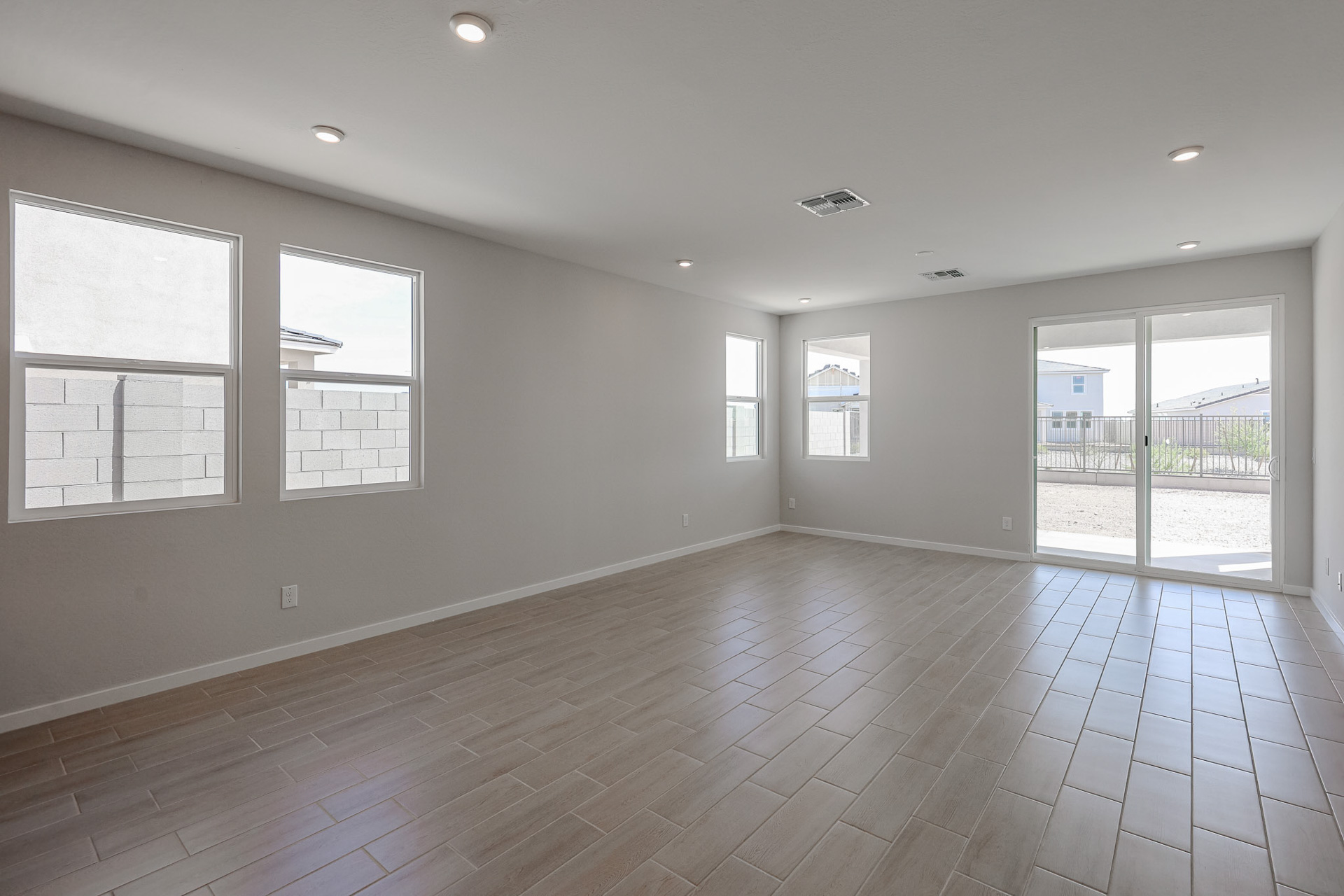 Empty room with light-colored tile floor, white walls, recessed ceiling lights, three windows on the left, and glass sliding doors on the right opening to a fenced backyard.