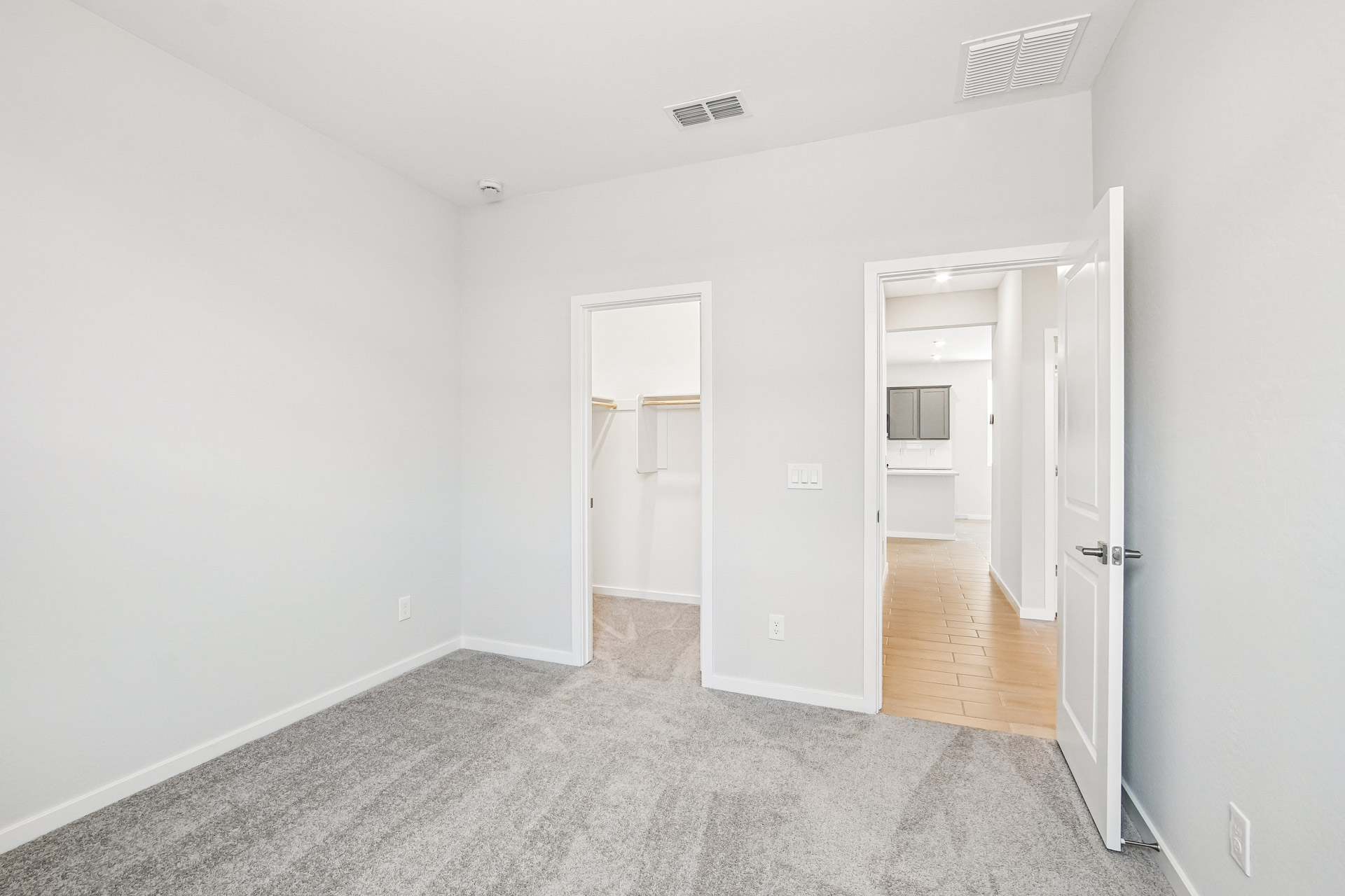 Empty room with light gray carpet, white walls, and two open doors leading to a closet and a hallway with wood flooring. Ceiling vents and an electrical outlet are visible.