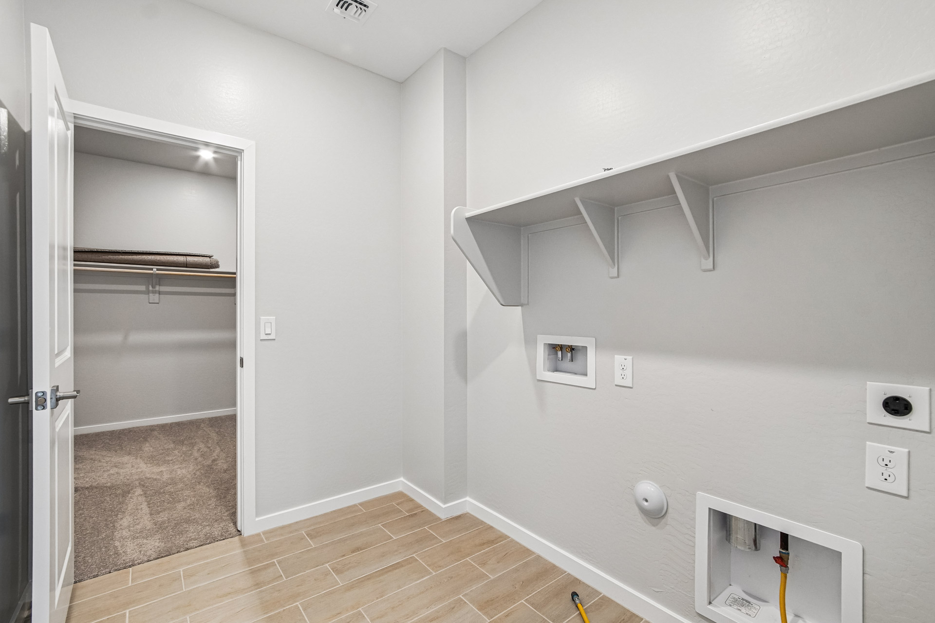 Laundry room with tile flooring, wall shelves, washer and dryer hookups, and an open door leading to a carpeted walk-in closet with shelving.