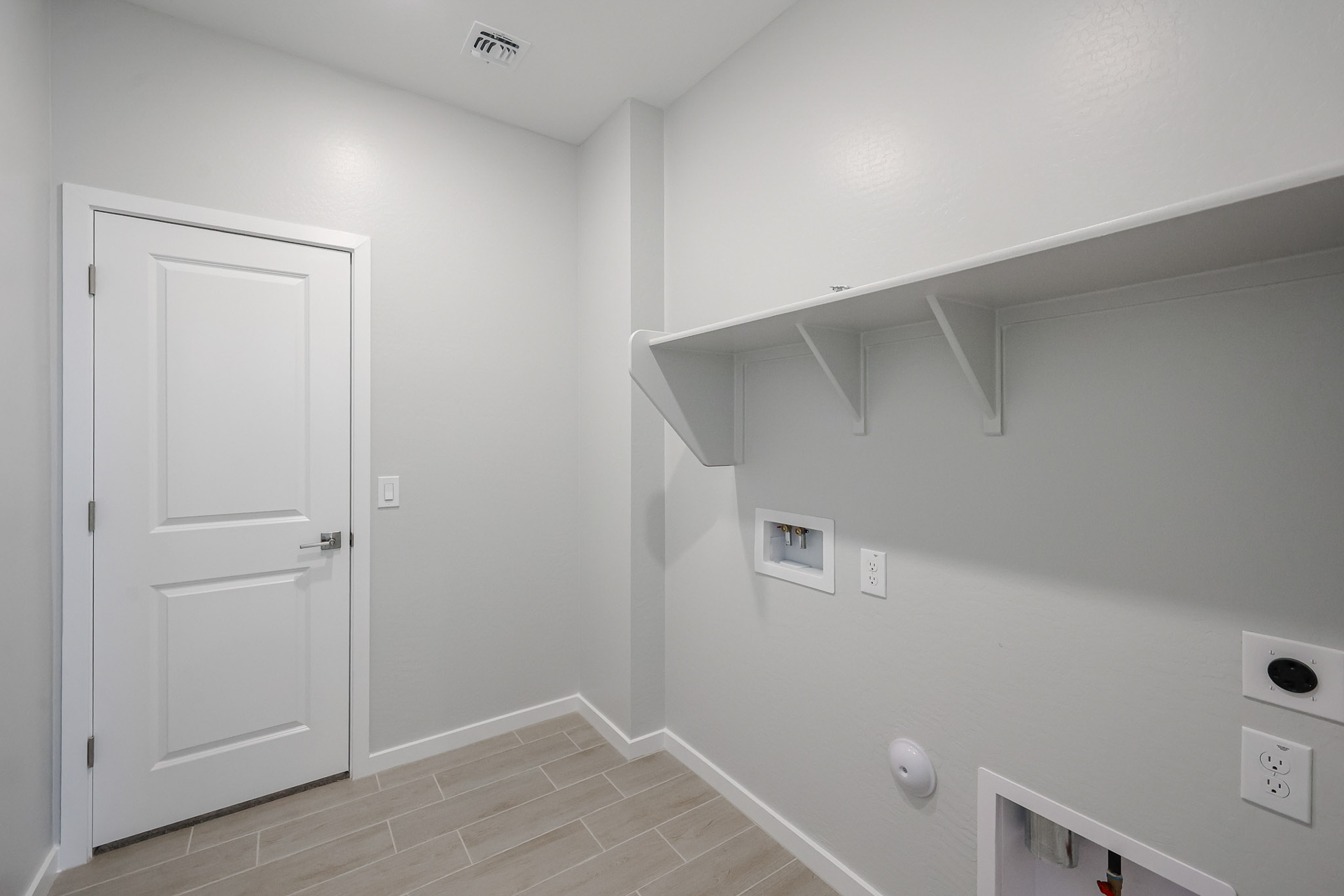Empty laundry room with white walls, tile floor, built-in shelf, washer and dryer hookups, and a closed white door.
