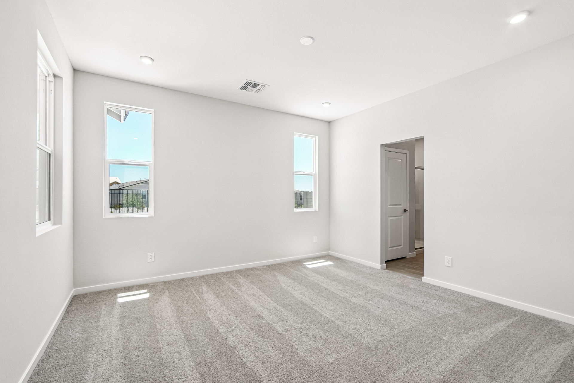 Empty, carpeted room with three windows, recessed lights in the ceiling, white walls, and an open doorway leading to another area. Natural light brightens the space.