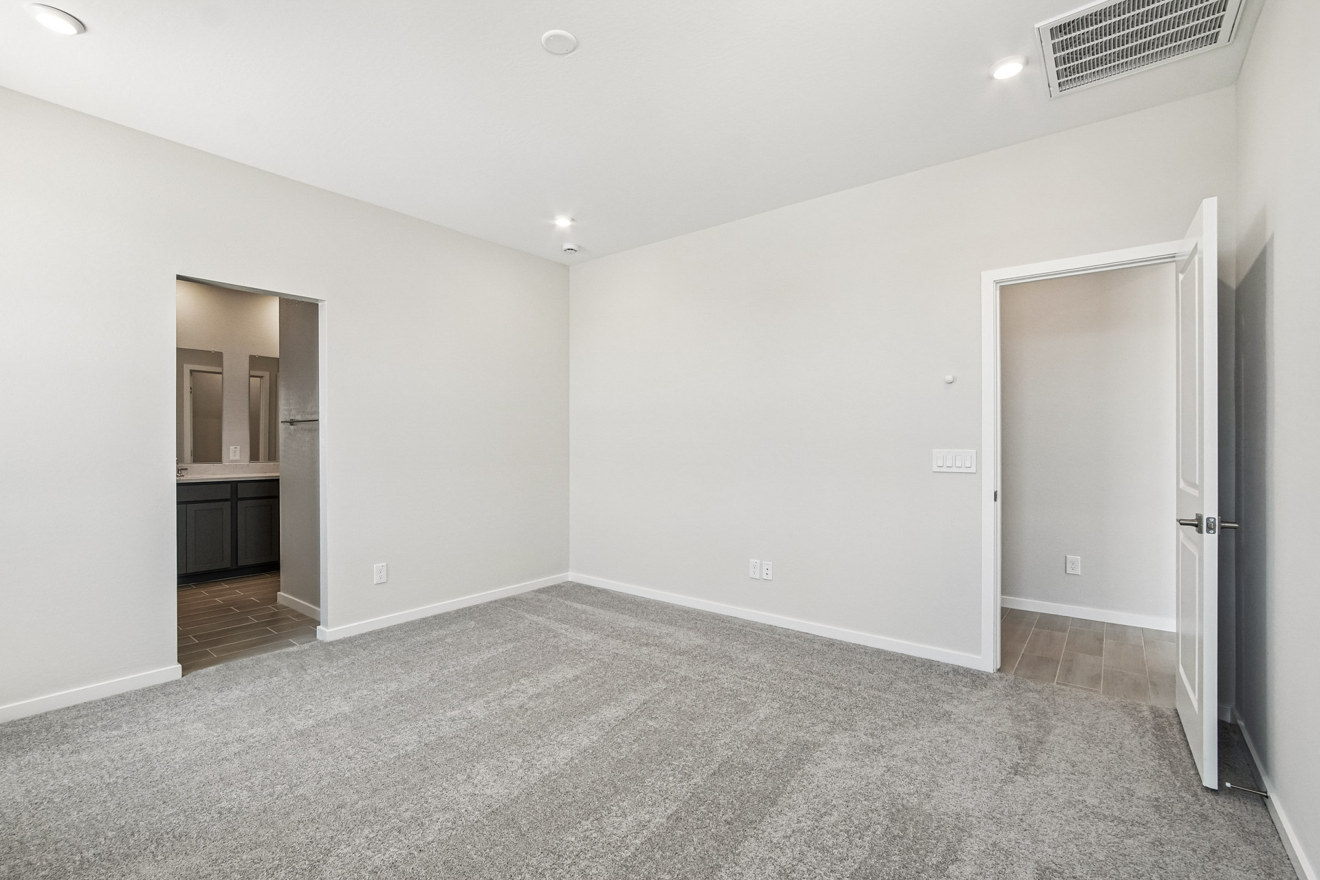Empty carpeted room with white walls, recessed lighting, and two open doorways; one leads to a bathroom, the other to a hallway.