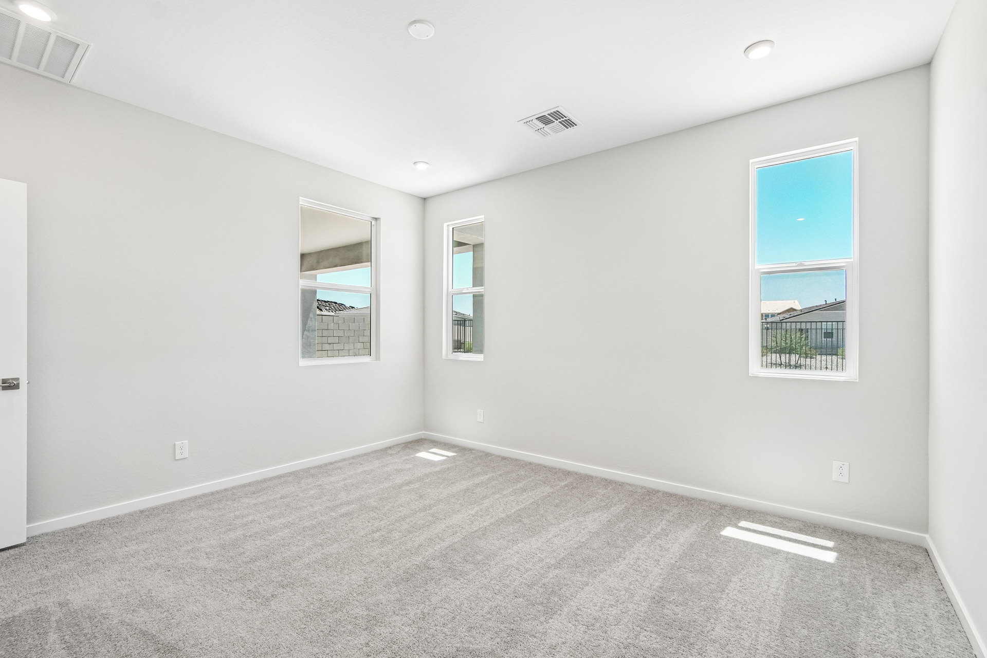 Empty, bright room with light gray carpet, white walls, two tall windows, and a door partially visible on the left. Ceiling lights are on, and daylight is streaming through the windows.