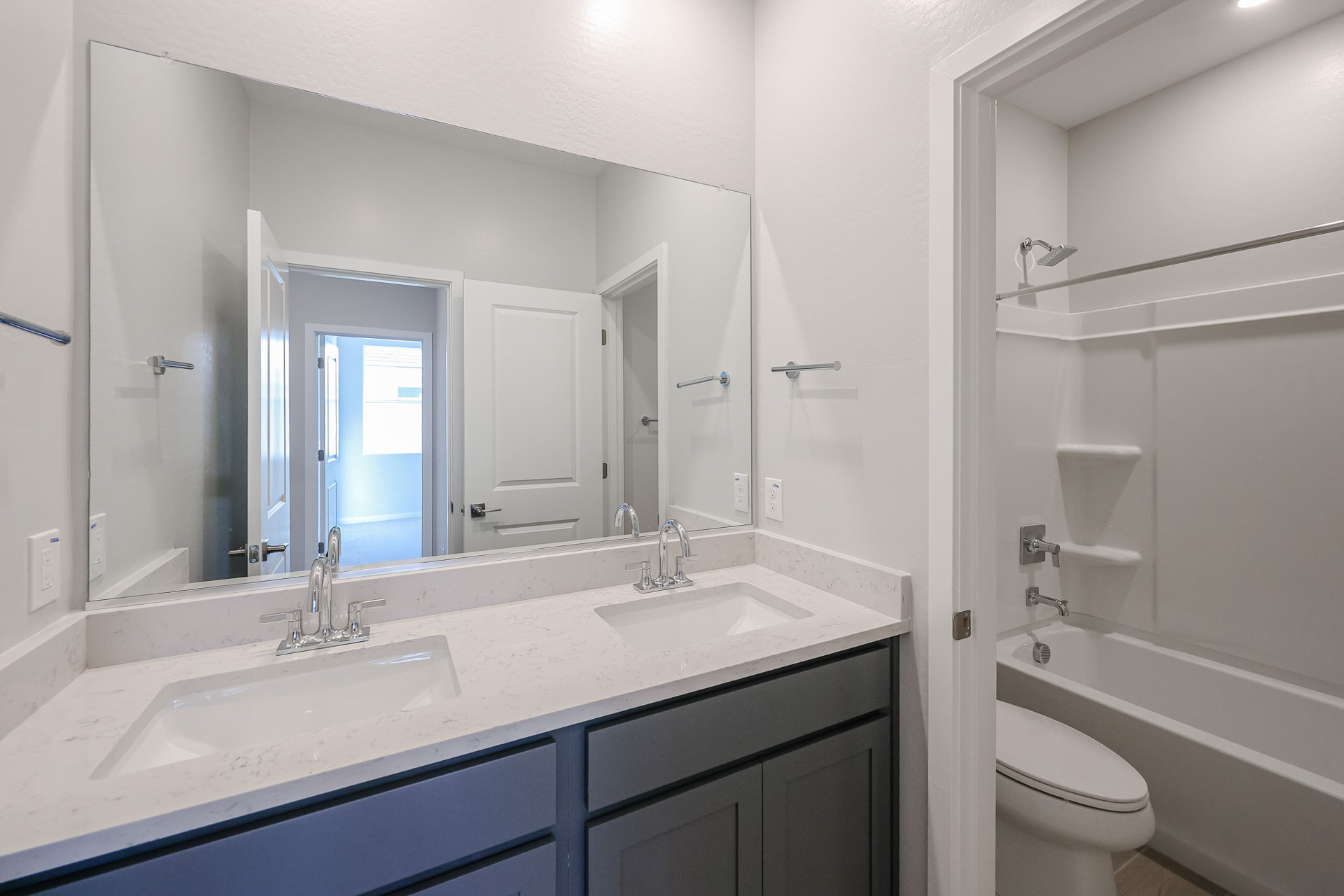 Modern bathroom with double sinks, large mirror, and view into a separate area with a bathtub, toilet, and shelves. Walls and countertop are light-colored; cabinets are dark.