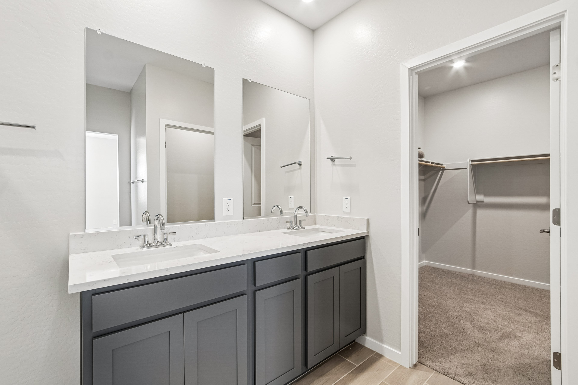 Modern bathroom with double vanity, gray cabinets, two mirrors, and an adjacent walk-in closet with shelves and carpeted floor. Walls and counters are light-colored.