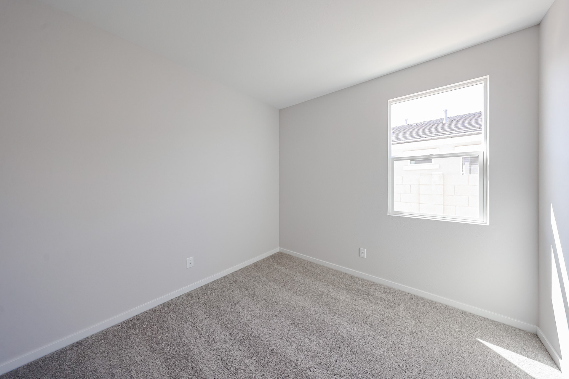 Empty, unfurnished room with light gray walls, beige carpet, a single window, and natural light coming in. The room appears clean and minimalistic.