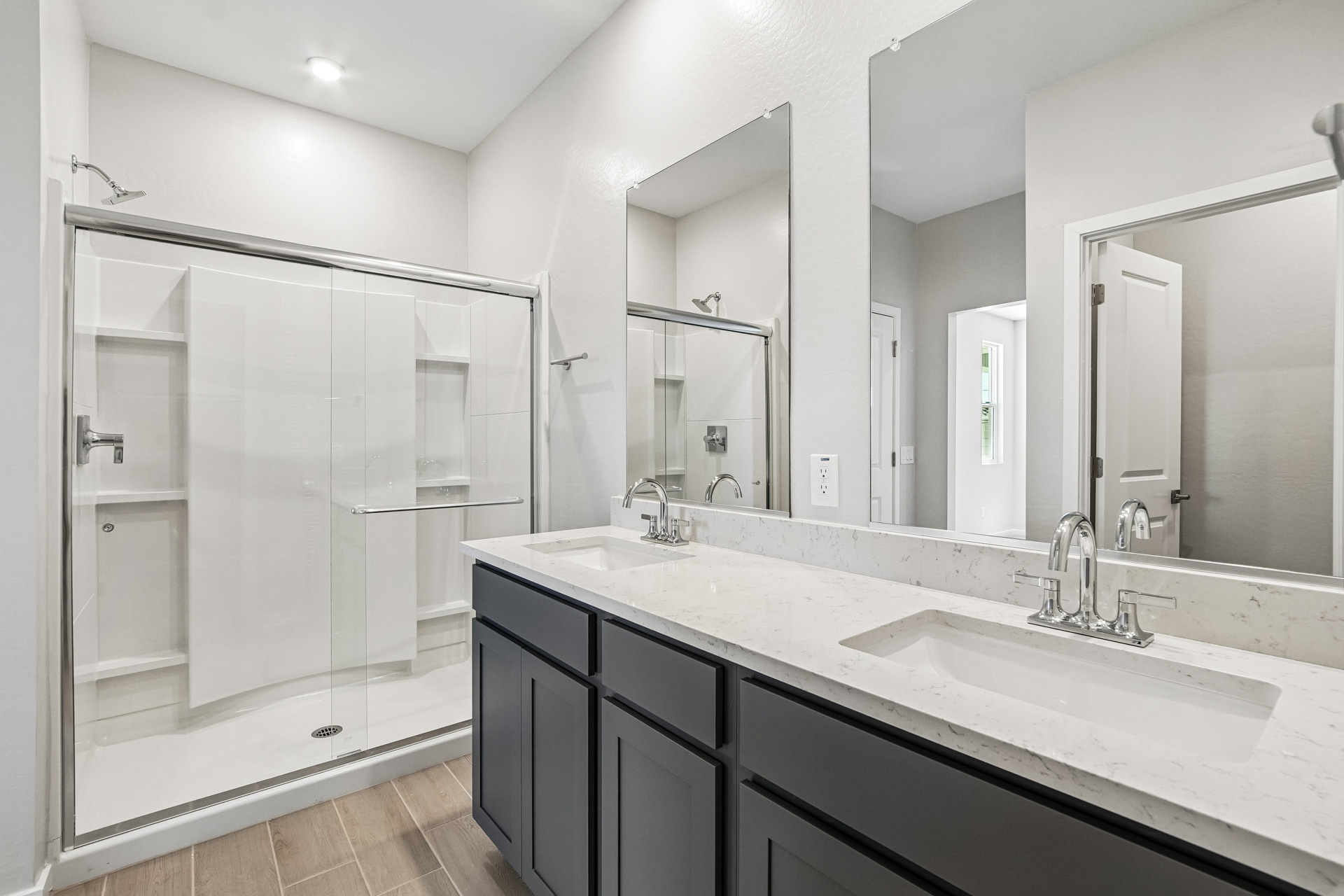 Modern bathroom with a double sink vanity, large mirrors, and a walk-in glass shower. Light walls, dark cabinets, and wood-look tile flooring create a clean, contemporary look.