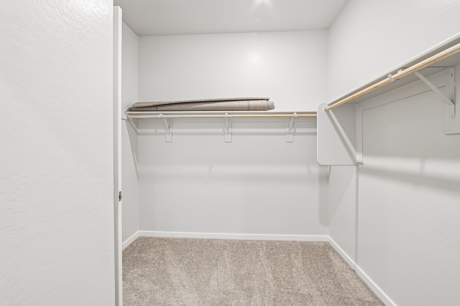 Empty walk-in closet with beige carpet, white walls, wooden hanging rods, and a rolled-up carpet piece resting on the top shelf.