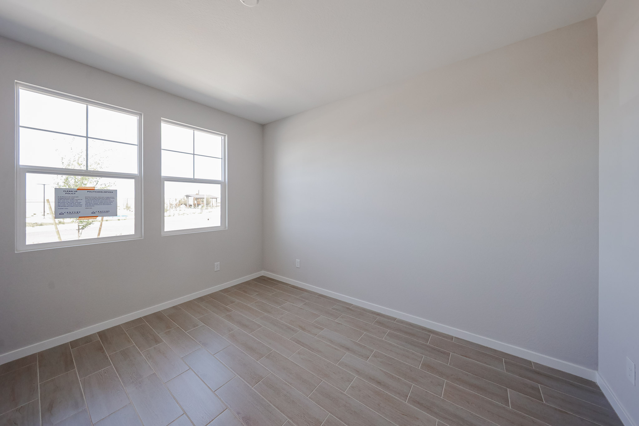 Empty room with light-colored walls, two large windows, and wood-look tile flooring. Sunlight enters through the windows, illuminating the clean, unfurnished space.