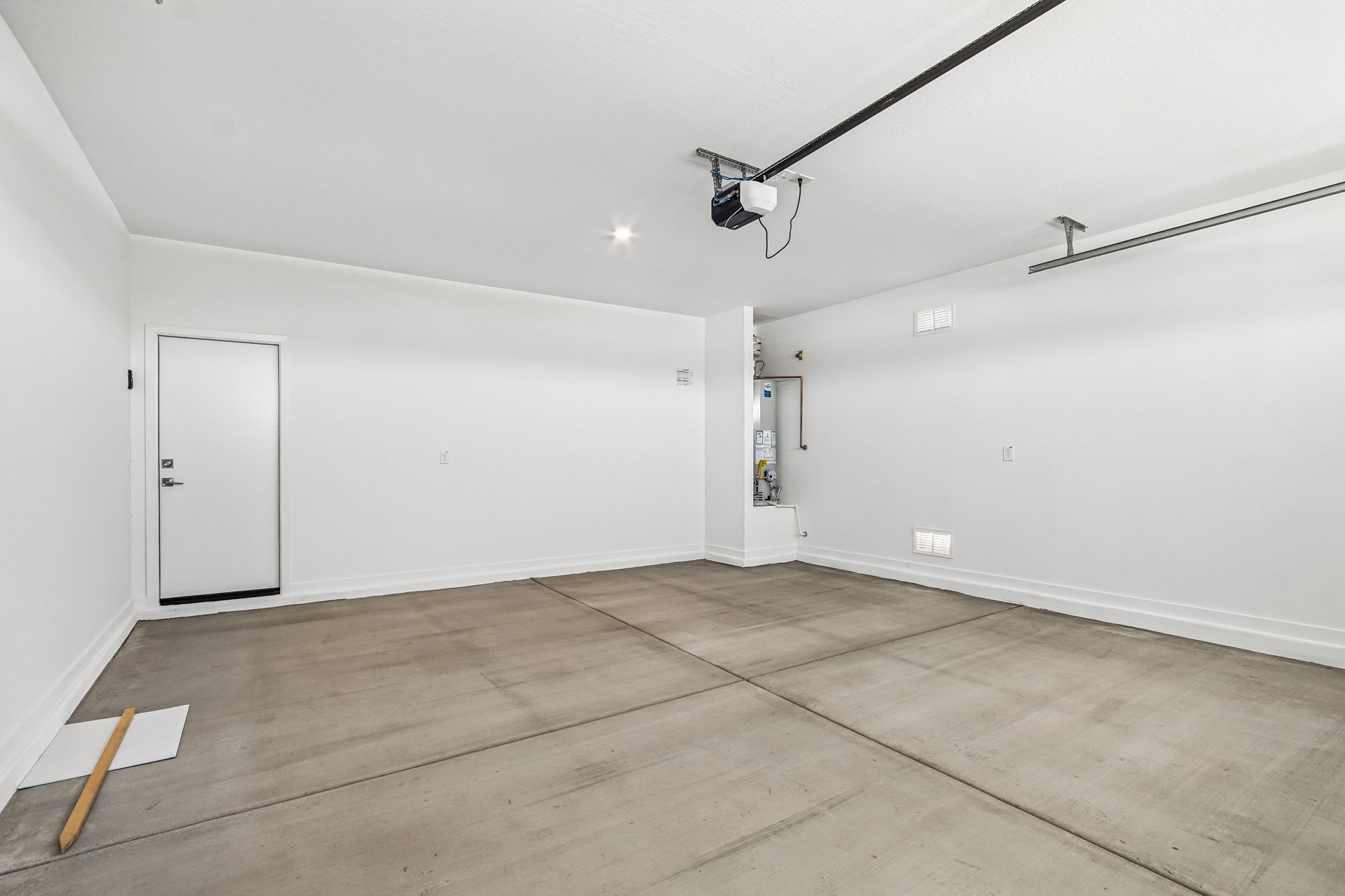 Empty two-car garage with white walls, concrete floor, ceiling-mounted garage door opener, and a side door on the left. A broom and a small sheet of paper are on the floor.