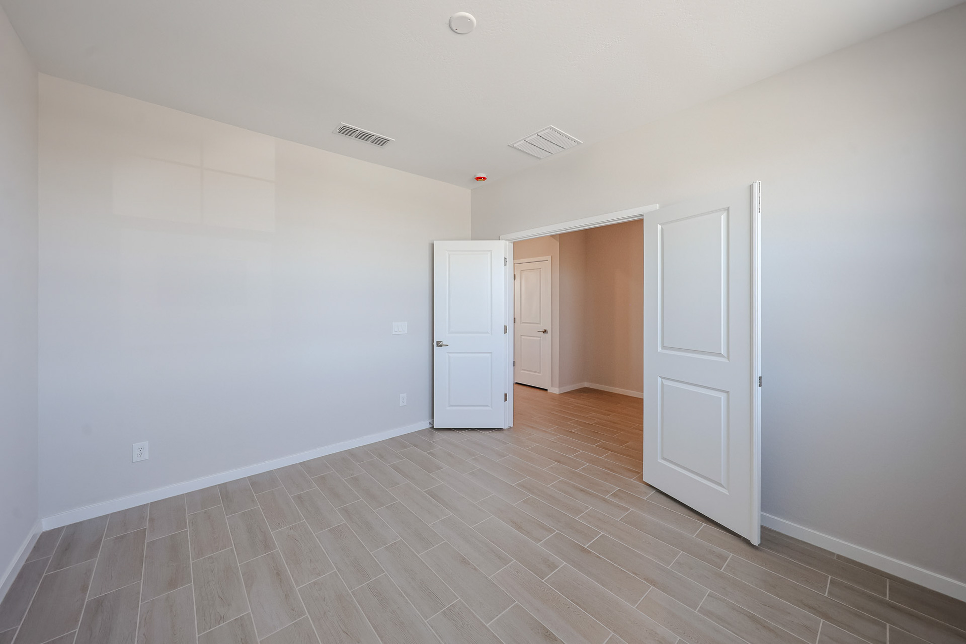 Empty room with beige tiled floor, white walls, and two open white doors leading to a hallway; ceiling vents and a red smoke detector are visible.