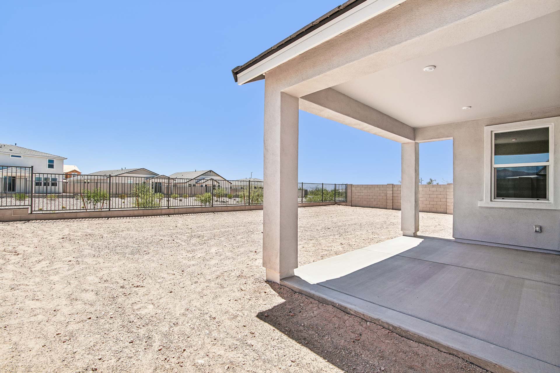 A covered patio extends from a modern house into a backyard with bare dirt and a metal fence, under a clear blue sky.