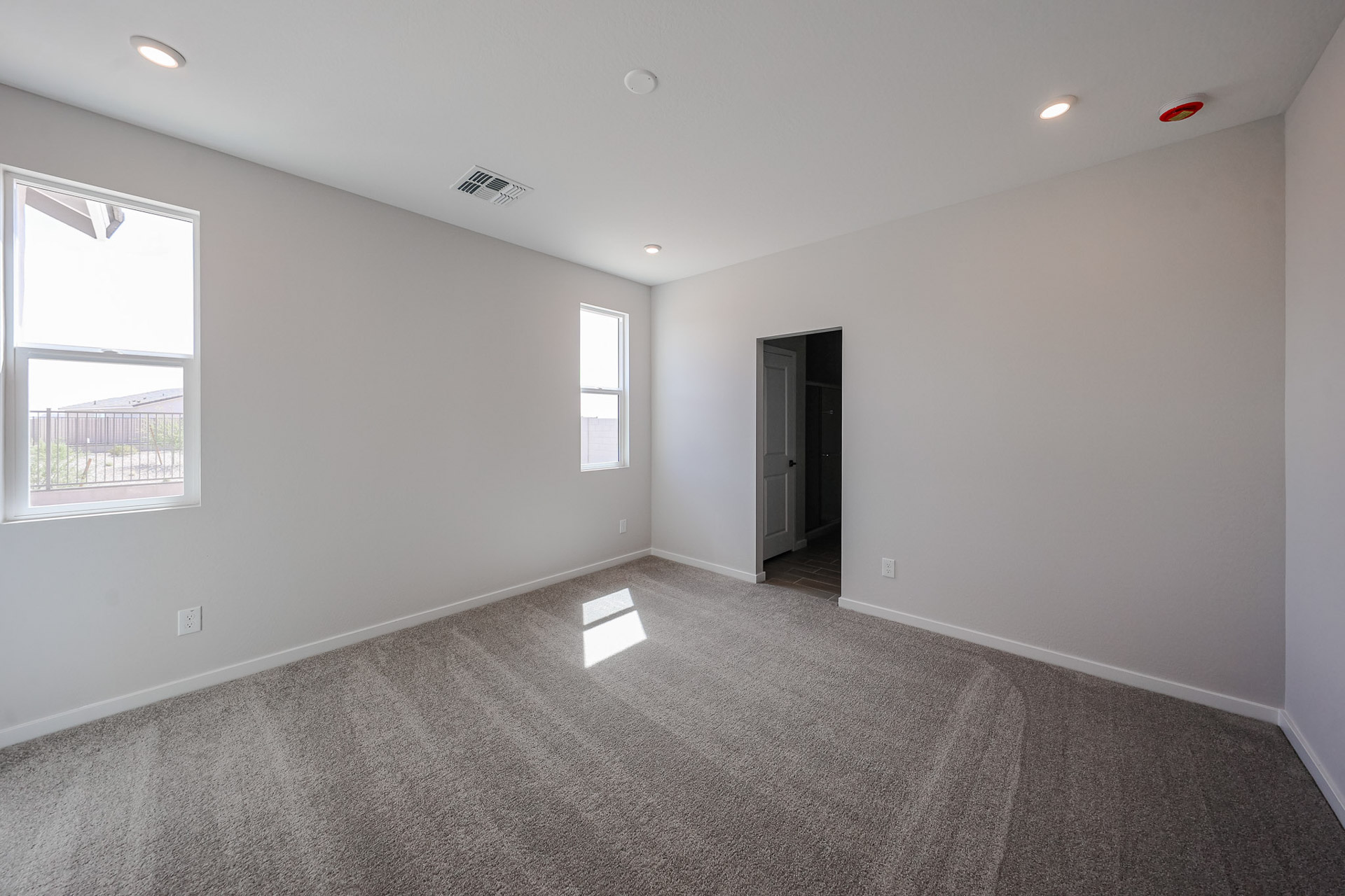 Empty room with light gray walls, two windows, beige carpet, and an open doorway leading to another area. Ceiling lights are recessed, and natural light enters from the windows.
