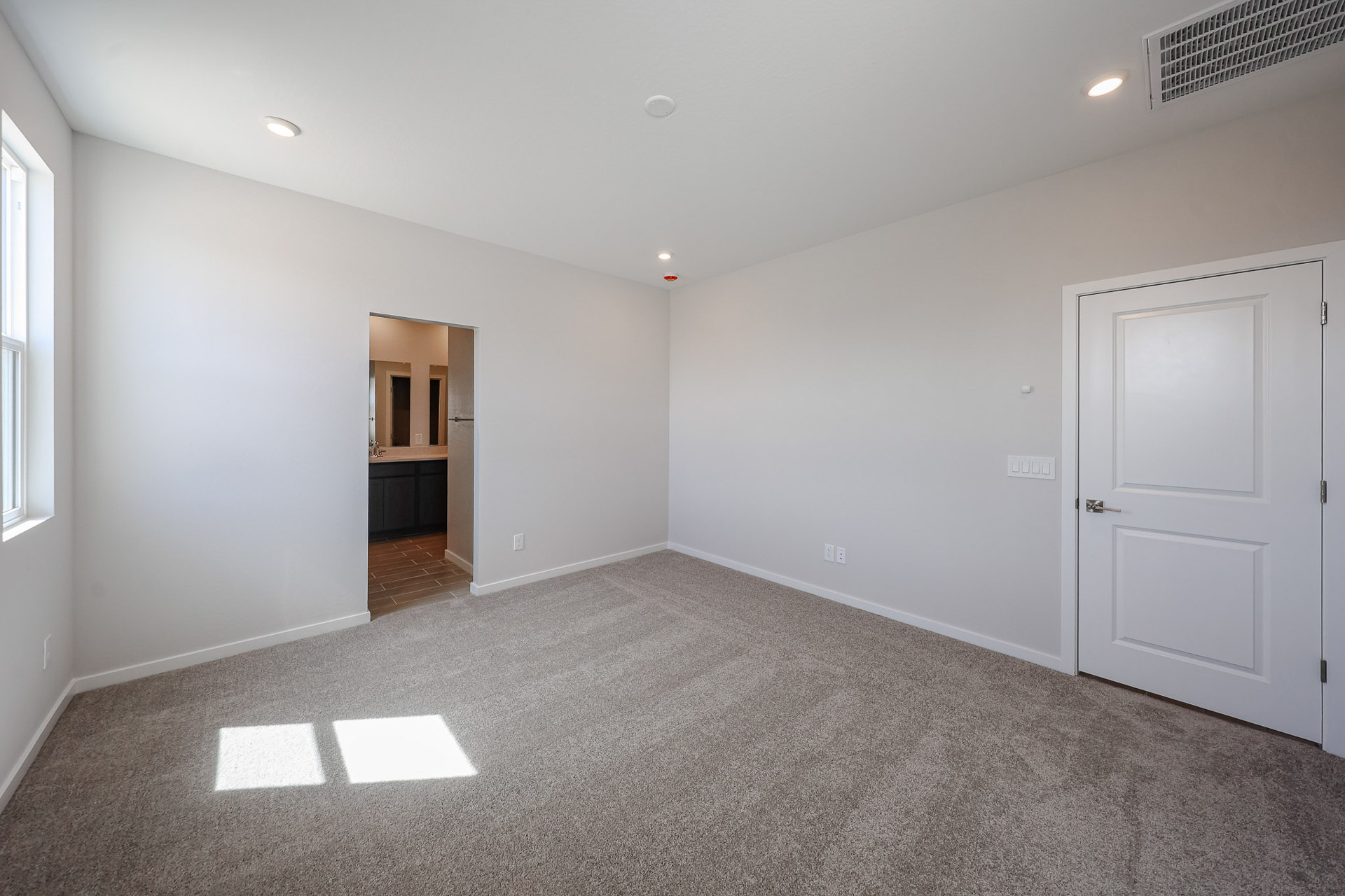 Empty carpeted room with white walls, a closed door, a window letting in sunlight, recessed ceiling lights, and an open doorway leading to a bathroom with a double-sink vanity.
