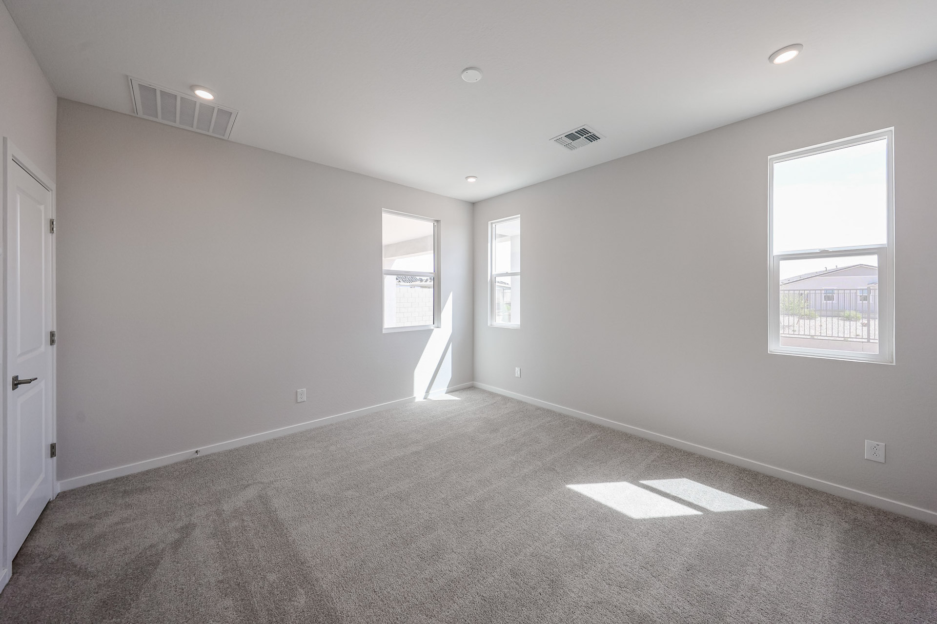 Empty, unfurnished room with light gray carpet, white walls, three windows, a closed white door, and recessed ceiling lights; sunlight enters through the windows.