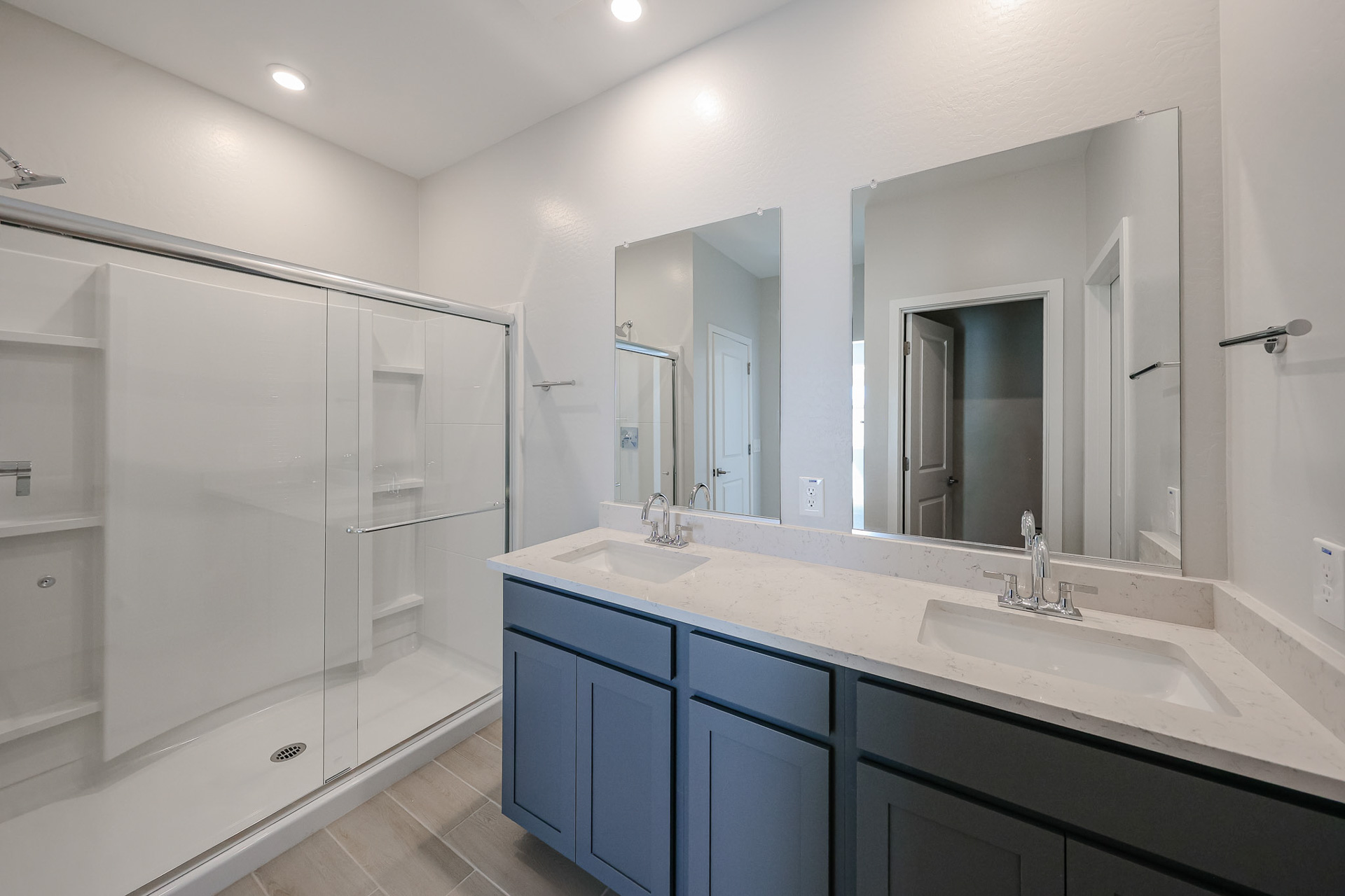 Modern bathroom with a glass-enclosed shower, double sink vanity with blue cabinets, large mirrors, and light-colored tile flooring.