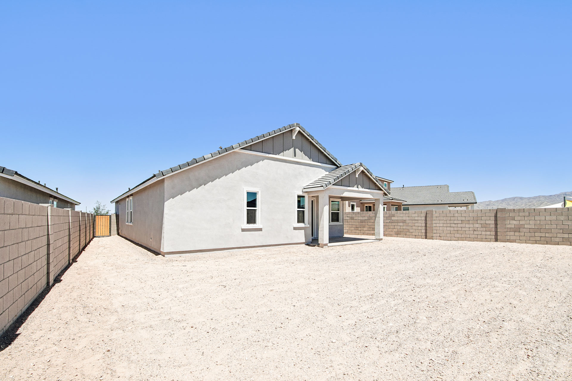 Single-story house with a small covered patio, light-colored exterior, and a large gravel backyard enclosed by a block wall under a clear blue sky.