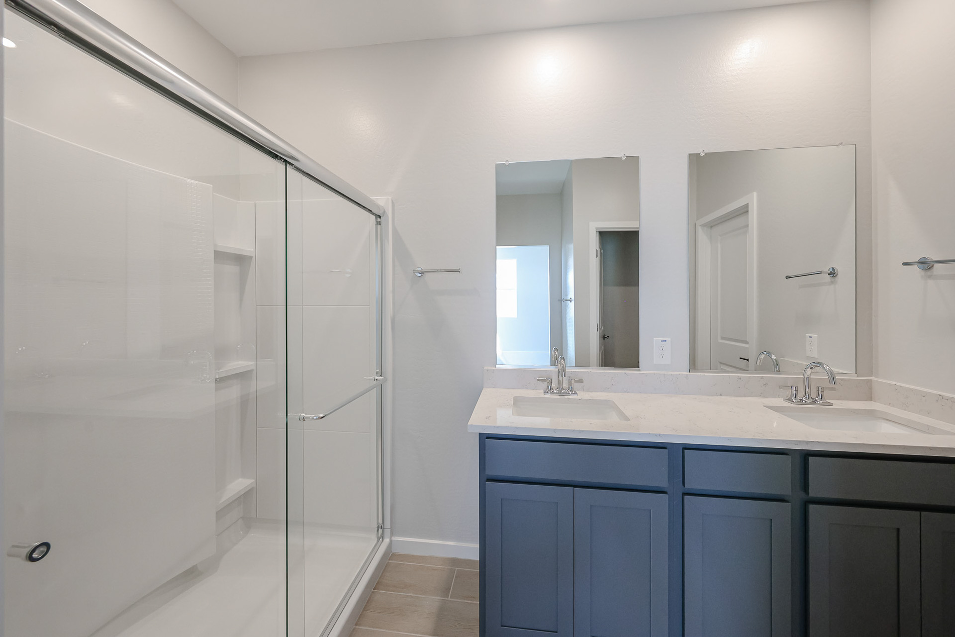 Modern bathroom with a glass-enclosed shower, double sinks with a marble countertop, blue cabinetry, two mirrors, and neutral-colored walls and flooring.