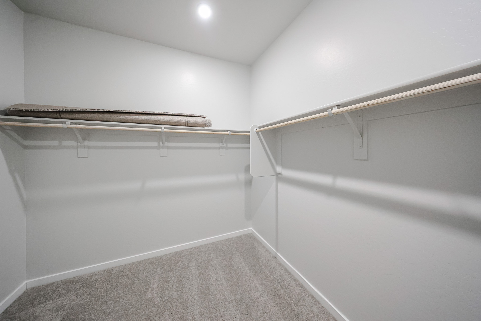 Empty walk-in closet with light gray carpet, white walls, overhead lighting, and two wooden shelves; a folded mattress or cushion sits on the left shelf.