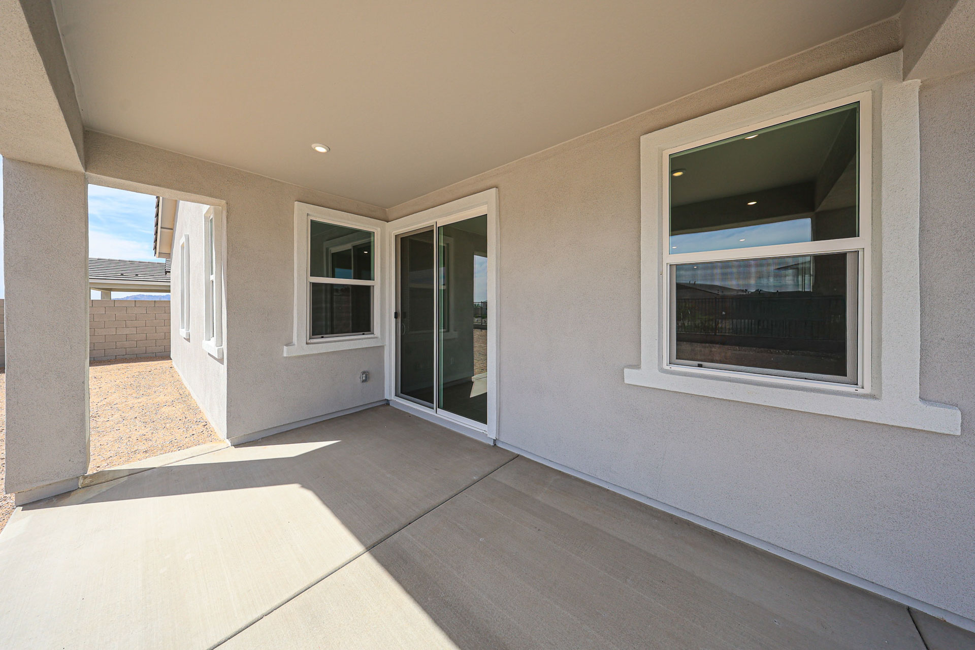 Covered patio area with beige walls, concrete floor, and two large windows next to a sliding glass door, leading to the side yard with gravel and a block wall.