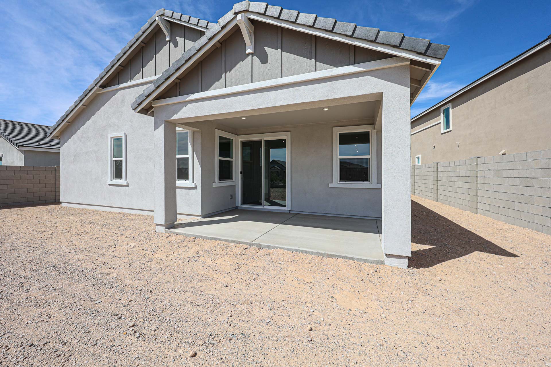 A modern single-story house with a covered patio, concrete slab, and a backyard of bare dirt, enclosed by gray cinder block walls under a blue sky.