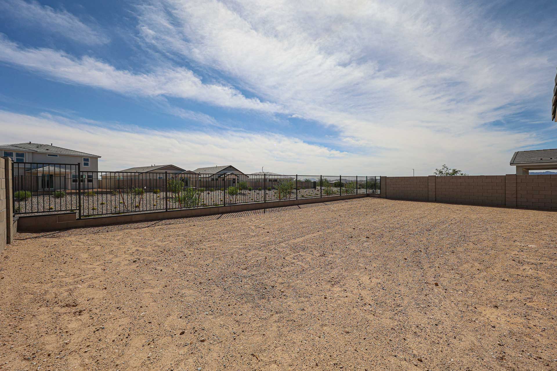 A fenced backyard with a gravel surface, bordered by a block wall and metal fence, under a partly cloudy sky.