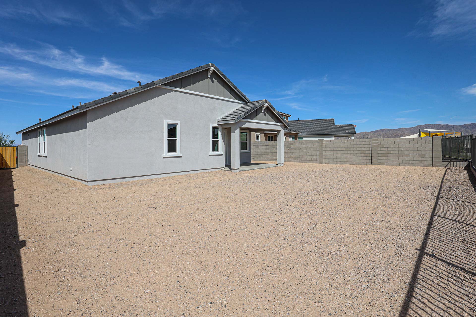 Single-story house with gray exterior, covered patio, and large, bare backyard with gravel ground, surrounded by a concrete block wall under a clear blue sky.