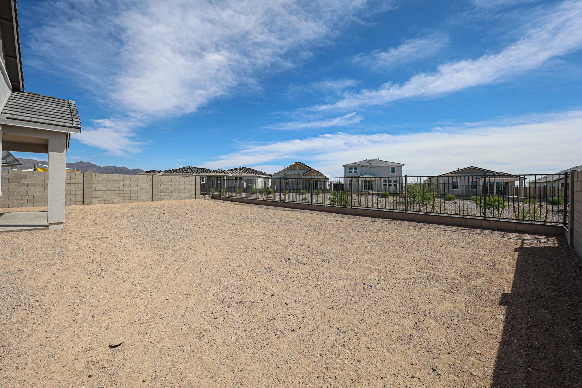 A large, fenced-in backyard with bare dirt ground, bordered by a block wall and metal fence, with neighboring houses and mountains in the background under a blue sky.