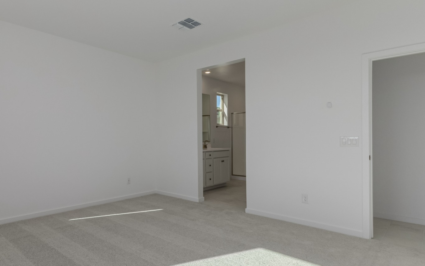 Empty room with light gray carpet, white walls, an open doorway leading to a bathroom with a vanity, and a closed door on the right. Ceiling vent and wall light switches are visible.