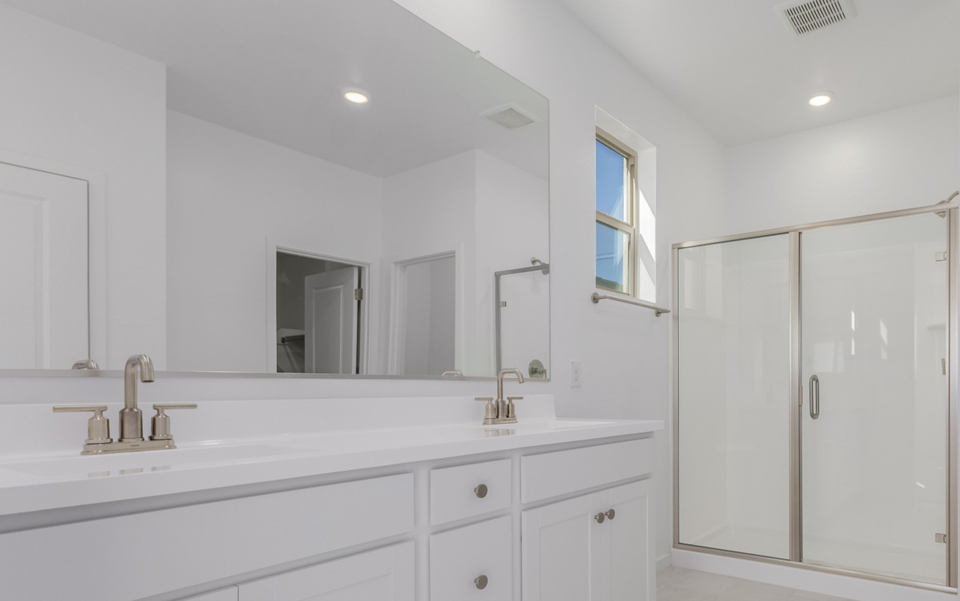 Modern bathroom with white cabinets, double sinks, large mirror, and a glass-enclosed shower. Walls and ceilings are painted white, and there is a small window above the shower area.
