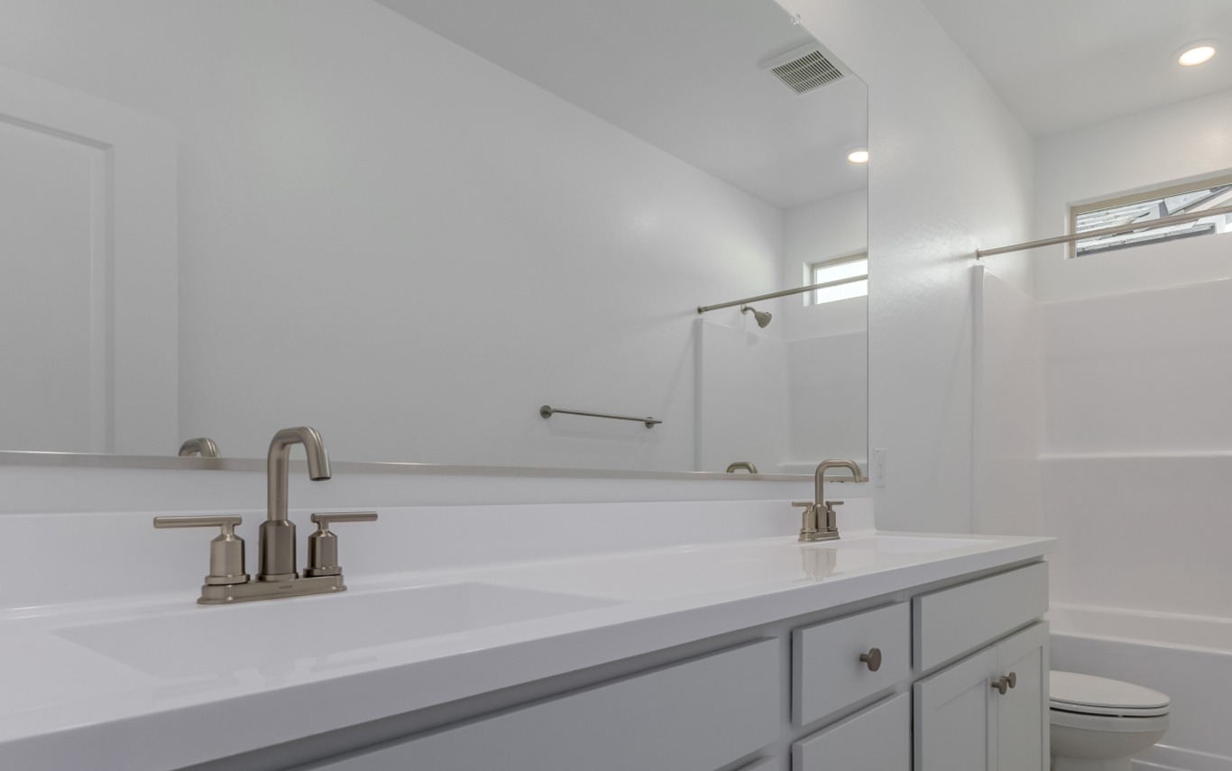 Modern bathroom with a double-sink vanity, large mirror, white countertop, silver fixtures, and a shower-tub combo in the background.