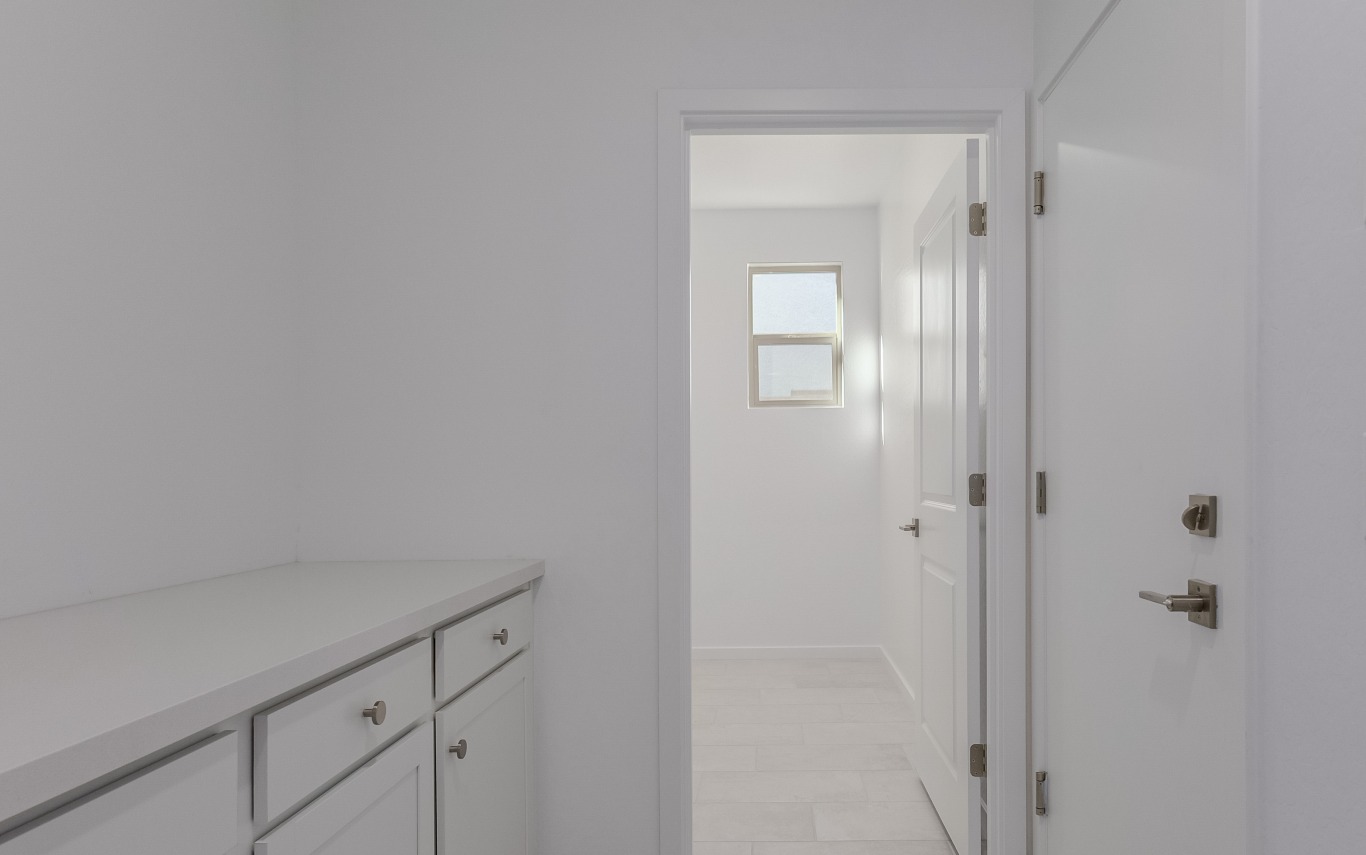A white hallway with cabinets on the left leads to a bright room with a window and a closed door, all featuring minimalist design.