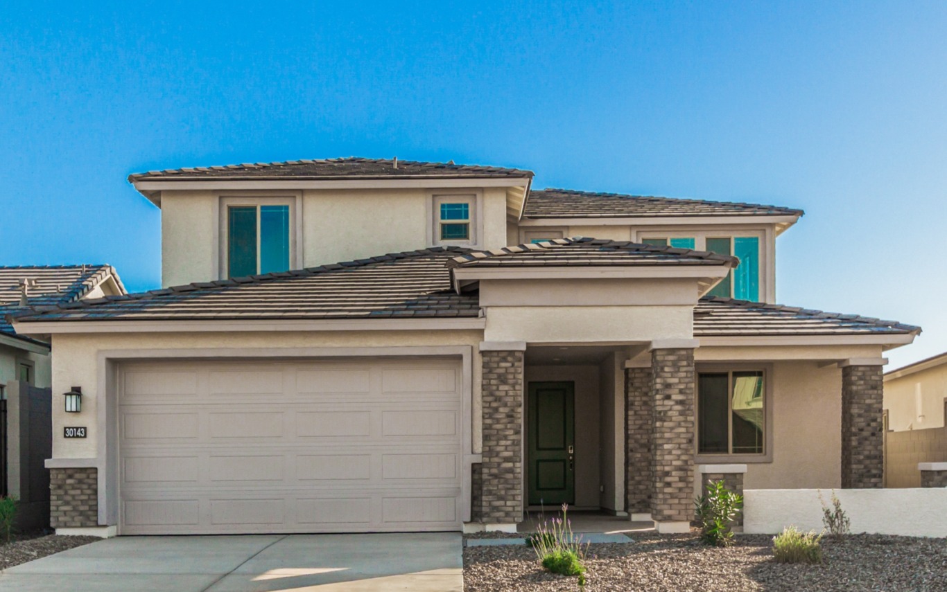Two-story modern suburban house with tan stucco exterior, green front door, double garage, and stone accents, set against a clear blue sky.