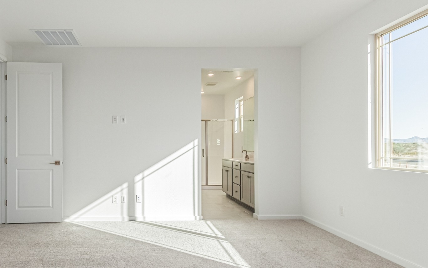 Bright, empty bedroom with beige carpet, white walls, and an open doorway leading to a bathroom with double sinks and a shower. Sunlight streams through a window on the right.