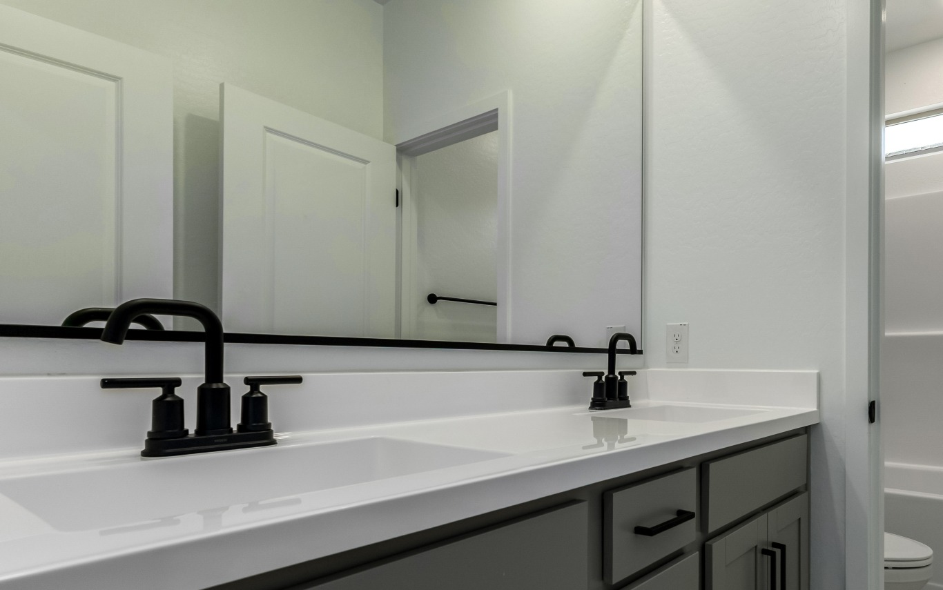 Modern bathroom with double sinks, black faucets, a large mirror, white countertop, gray cabinets, and open white door.