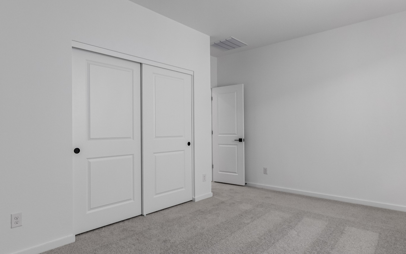 Empty room with beige carpet, white walls, a closed double sliding closet door, and an open single door leading to another space.