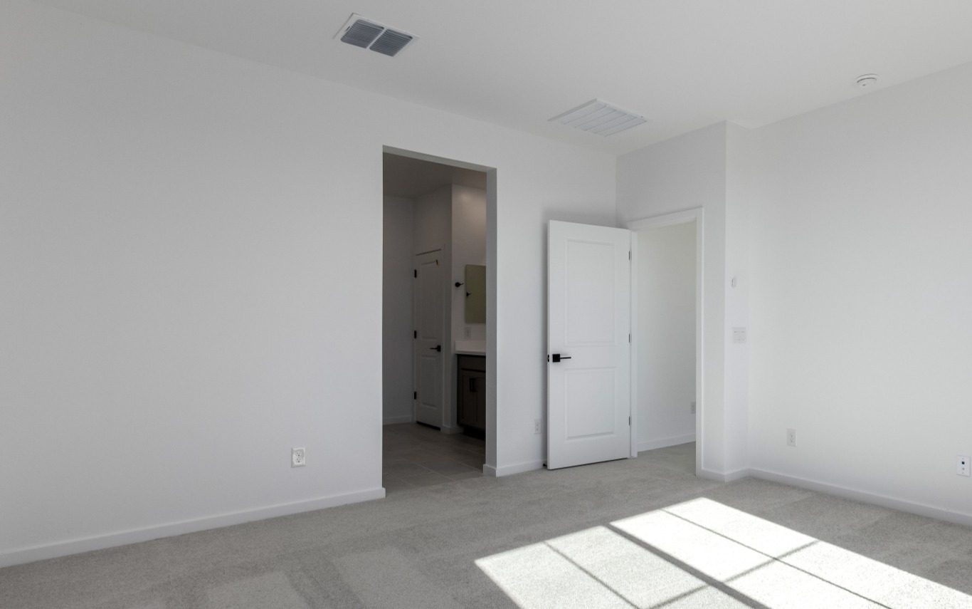 Empty room with light gray carpet, white walls, and an open door leading to a hallway and what appears to be a bathroom; sunlight streams through a window onto the floor.