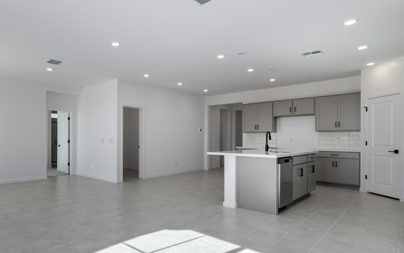 Open modern kitchen with gray cabinets, white countertops, an island, and tile flooring, connected to a spacious, empty living area with white walls and recessed lighting.