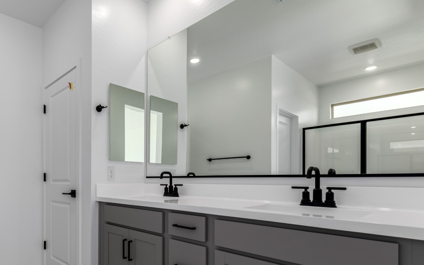 Modern bathroom with double sinks, black fixtures, large mirror, gray cabinets, glass shower enclosure, and white walls.