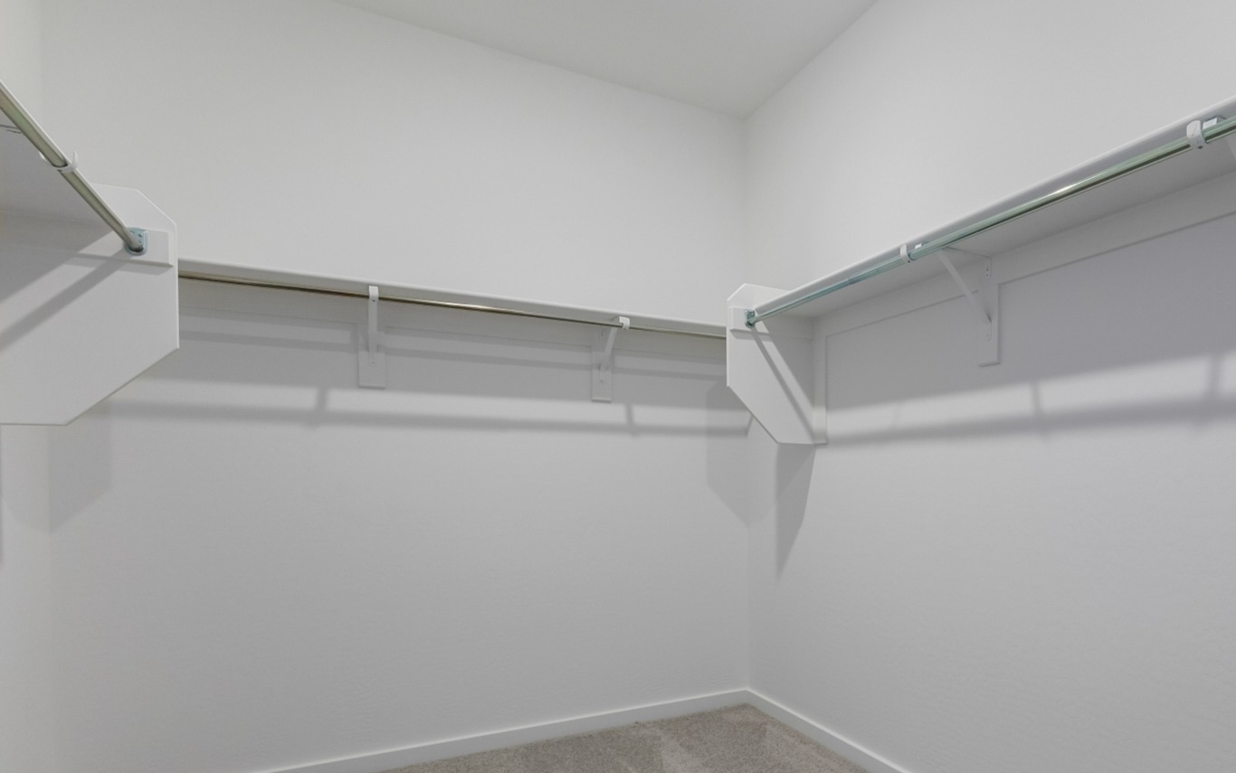 Empty walk-in closet with white walls, beige carpet flooring, and metal hanging rods mounted on two walls.