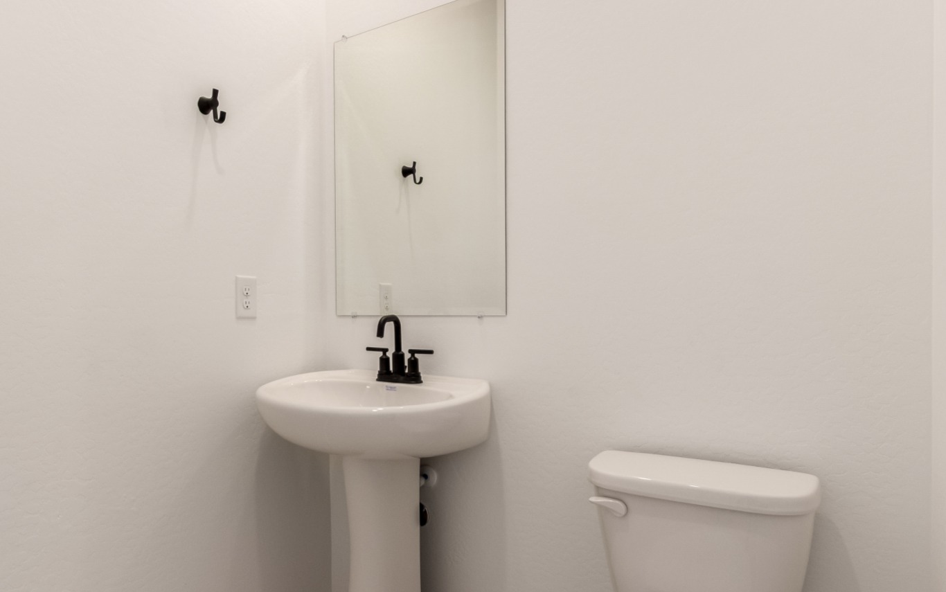 A small bathroom with a pedestal sink, rectangular mirror, toilet, two black wall hooks, and black faucet; white walls and minimal decor.