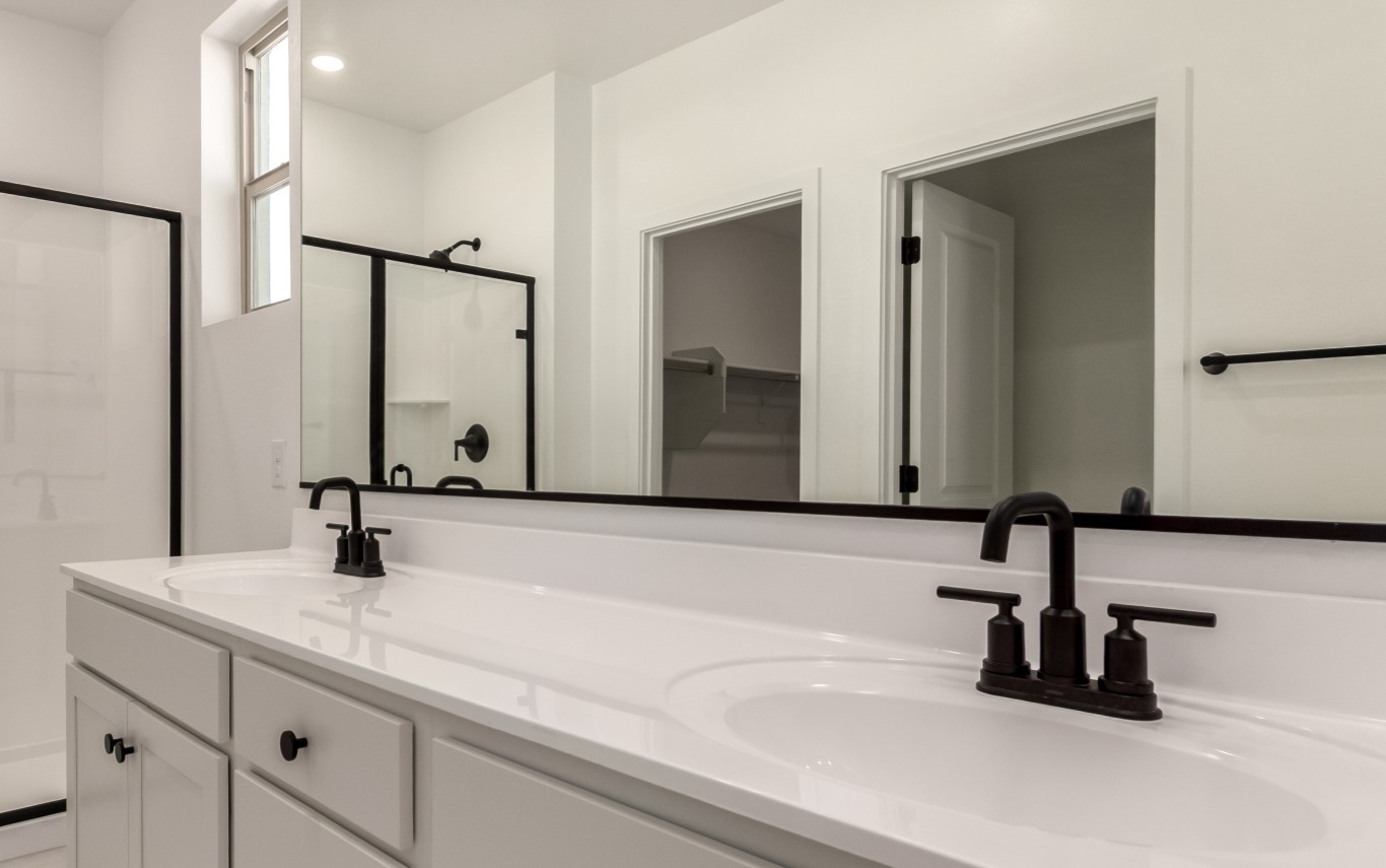 Modern bathroom with a double sink vanity, black faucets, a glass shower enclosure, white cabinets, and a walk-in closet visible through an open door.