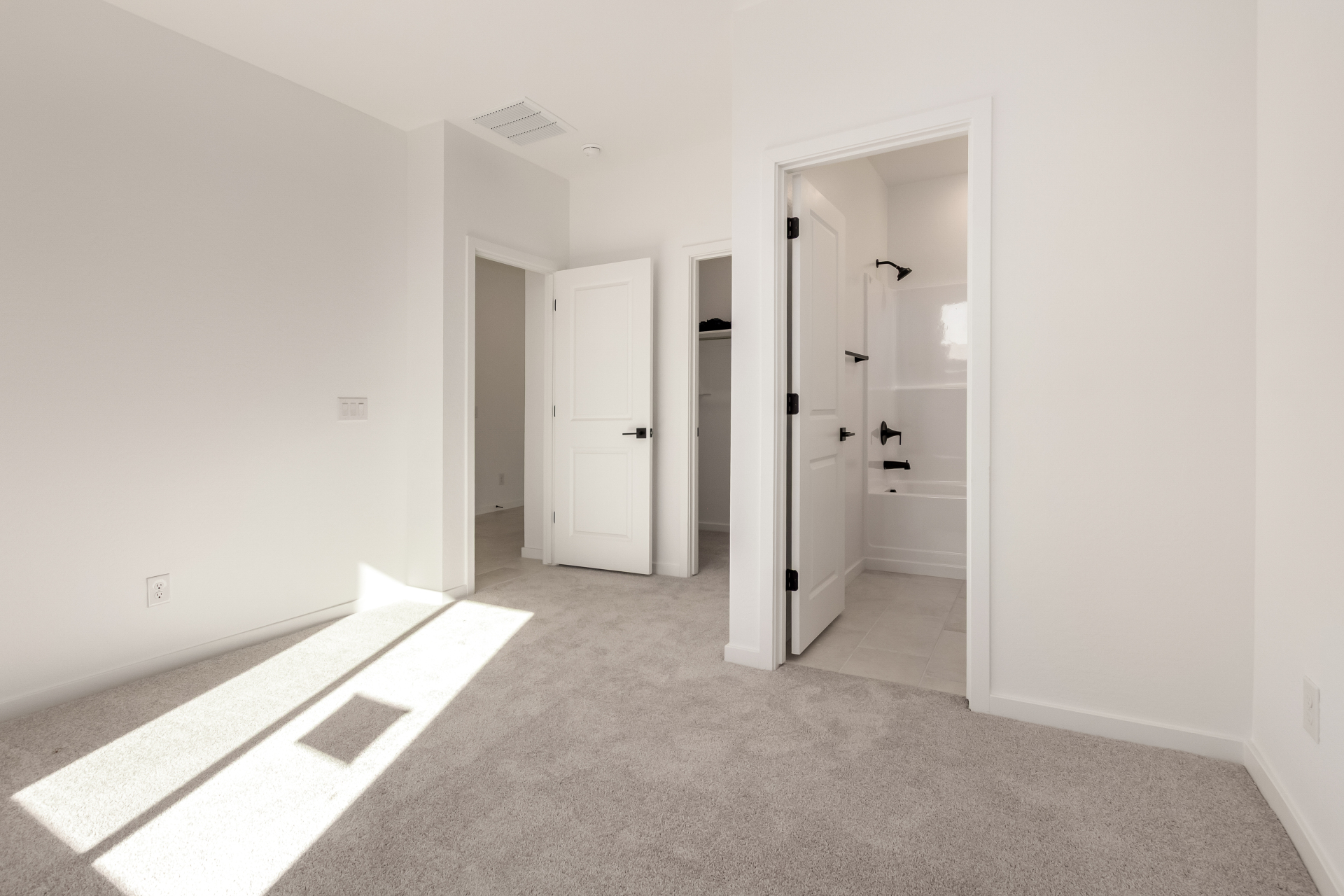 Empty bedroom with light gray carpet, white walls, open doorways to a closet and bathroom, and sunlight streaming through a window onto the floor.
