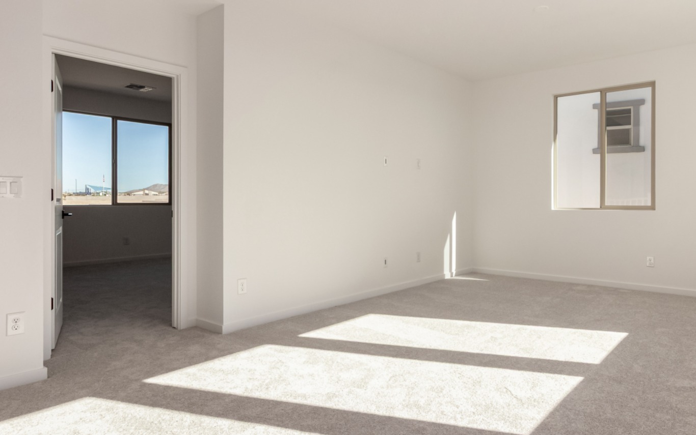 Empty, sunlit room with light carpet, white walls, and two windows; one wall has a door leading to an adjacent room.
