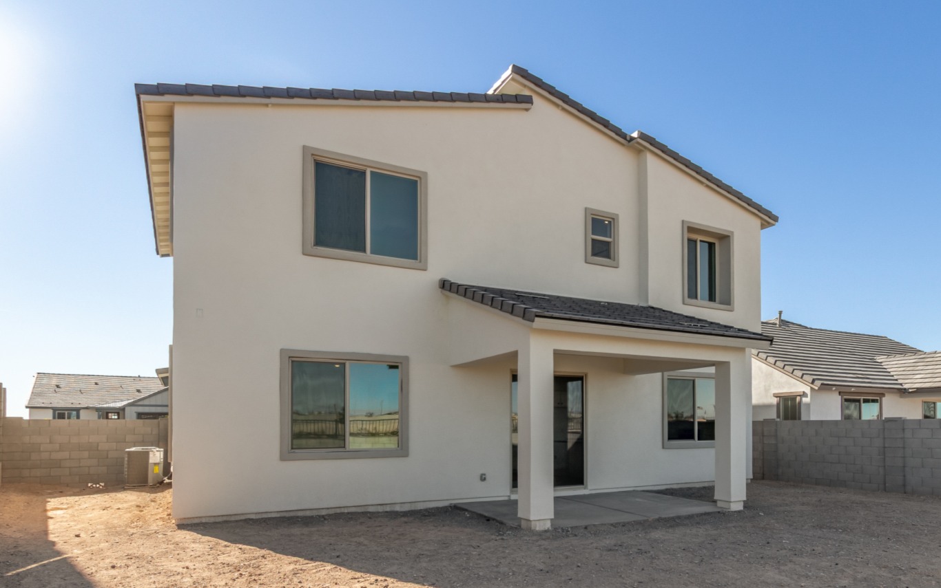 Two-story modern house with light-colored exterior, multiple windows, and a covered back patio, situated in a fenced yard with bare ground and clear sky.