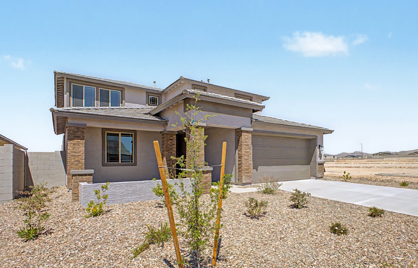 Two-story suburban house with a gray exterior, stone accents, and a two-car garage, surrounded by a desert landscape and small plants under a clear blue sky.